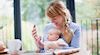 Auburn haired mother in blue cardigan showing young baby in white baby grow smartphone