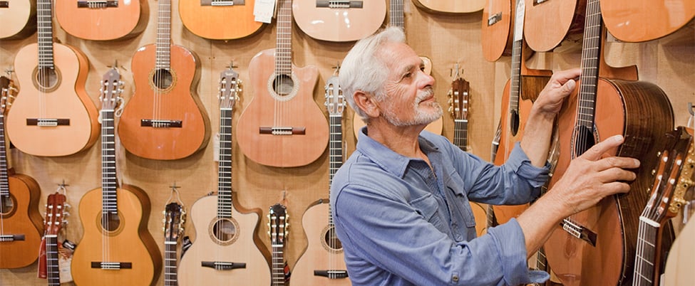 Grey haired and bearded man wearing blue flannel shirt looking at guitars hung on wall