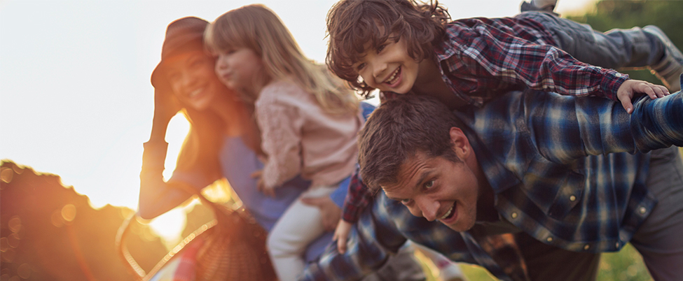 Family playing in a field, son climbs on fathers back while mother holds daughter in background