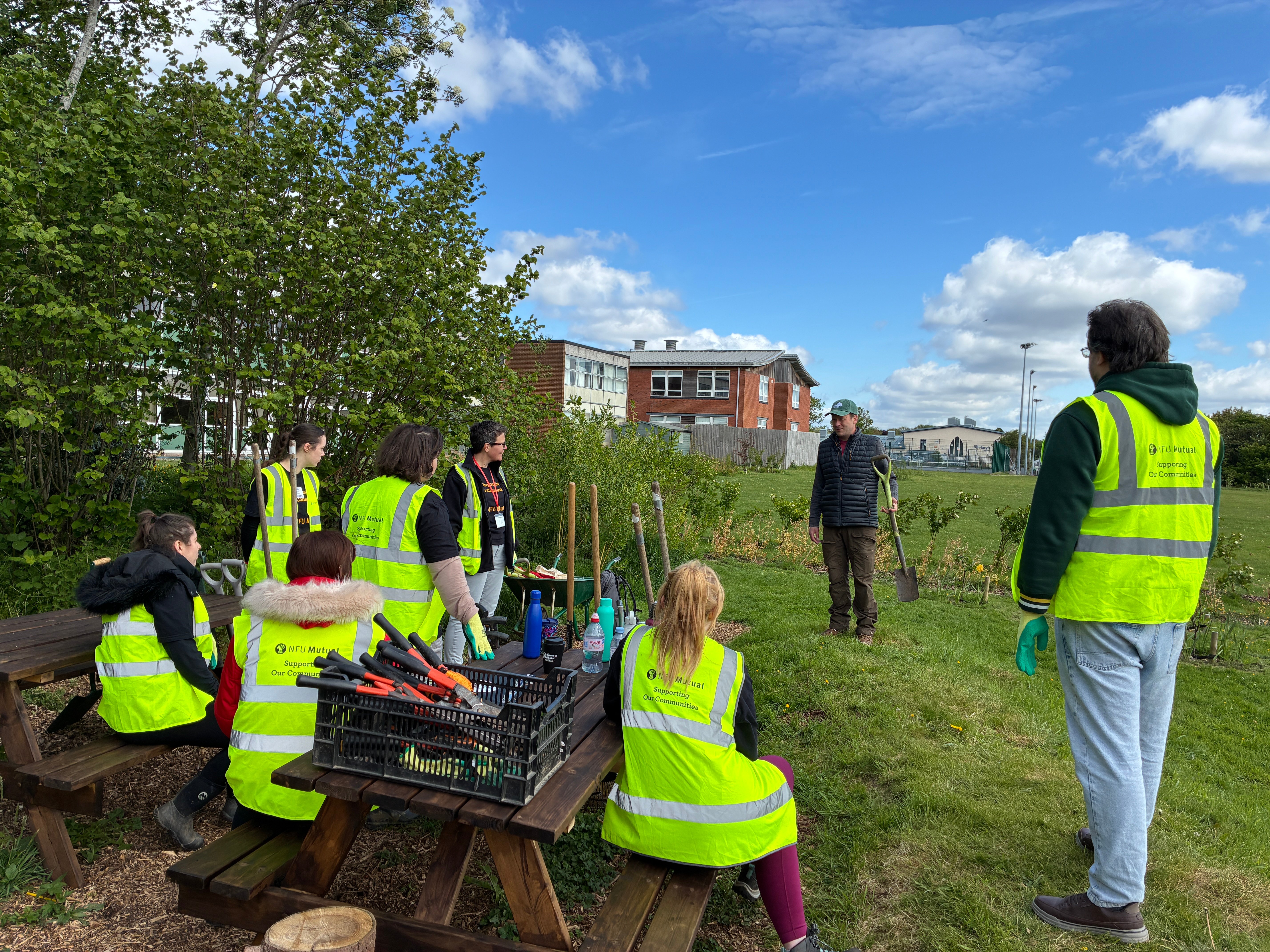 A group of people wearing yellow hi-vis jackets, looking at a man speaking while holding a shovel.