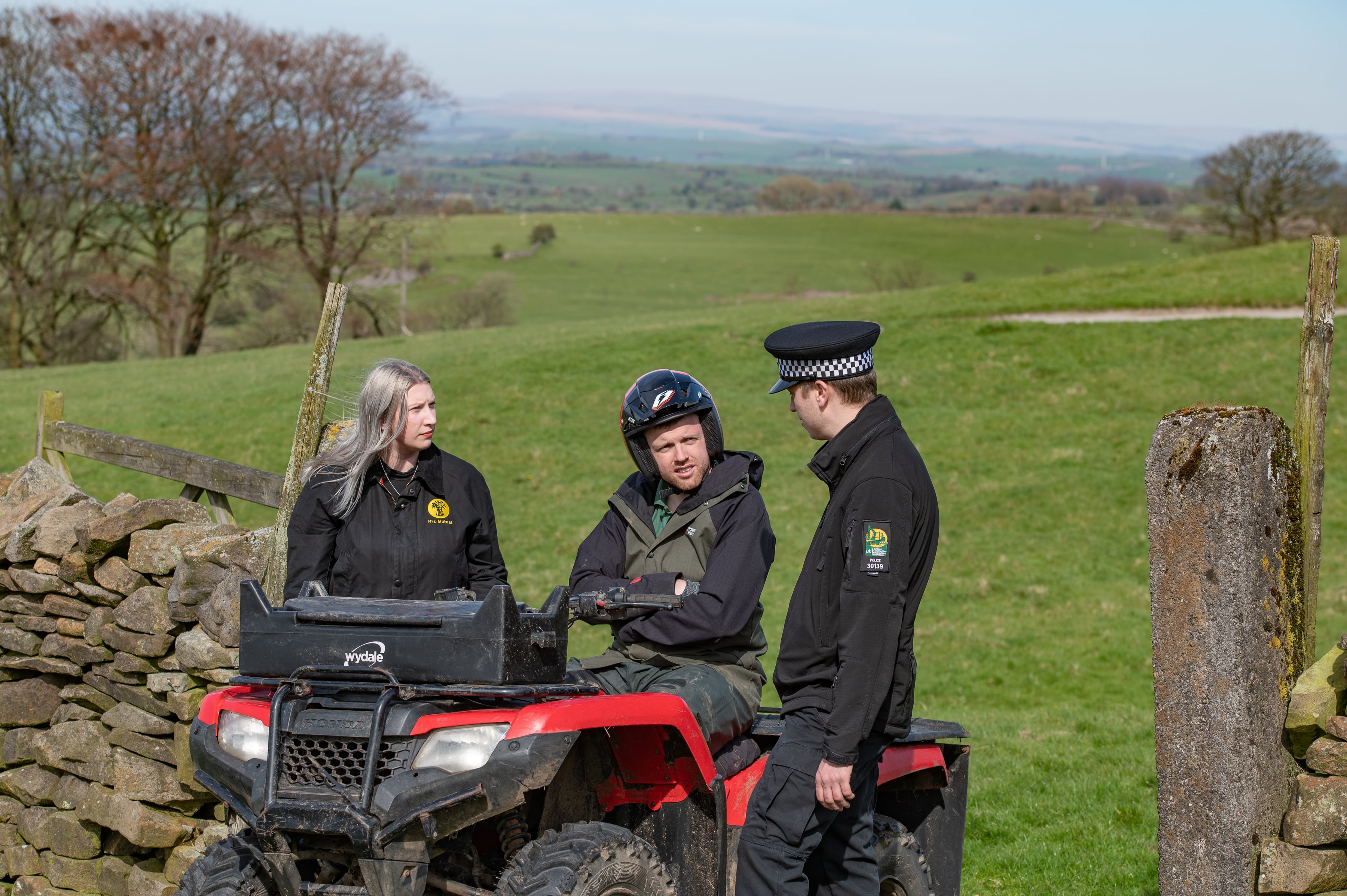 Three people in a field, one sat on a quad bike, one with an NFU Mutual coat on and one police officer.